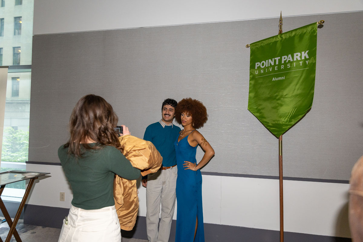 Two female students pose for a photo while another person takes the picture on their phone. 