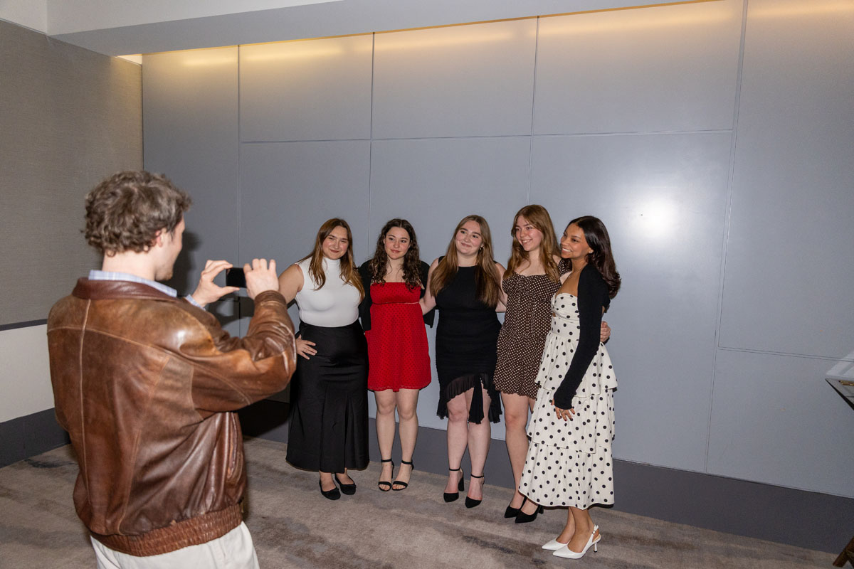 A group of students pose for a photo while a man takes their picture on a phone.